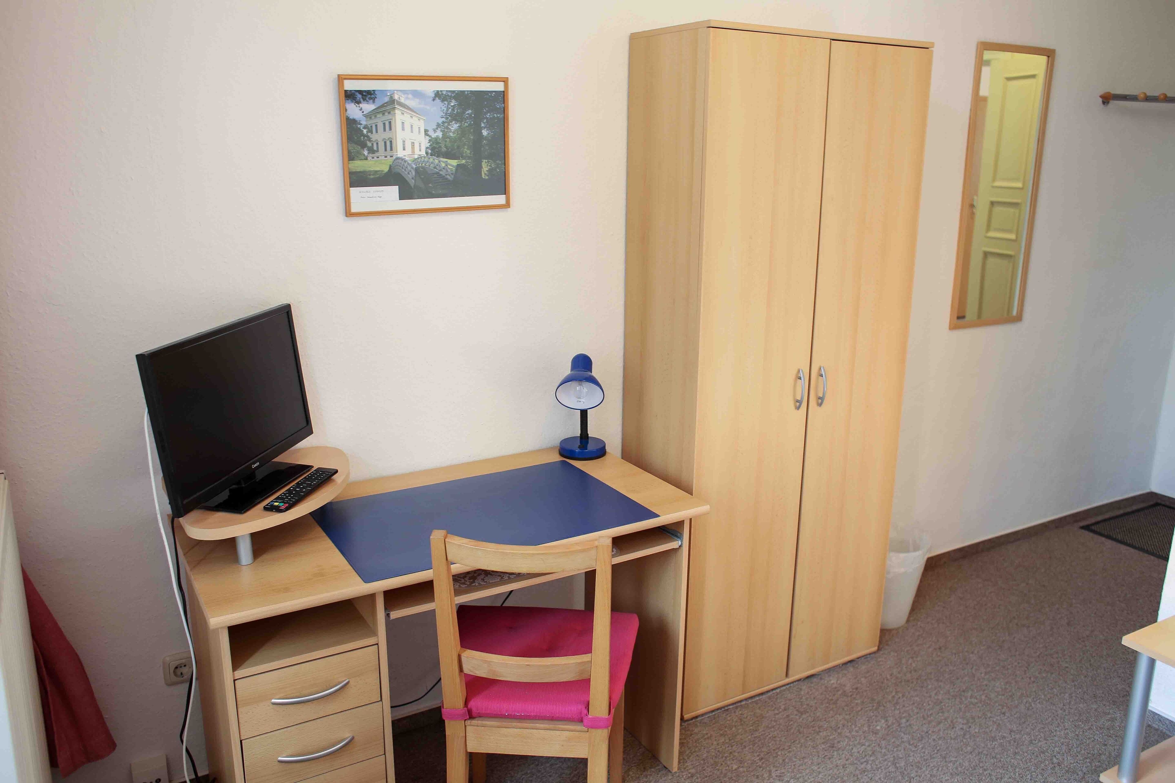 A room with a wooden desk, a chair, a television, a lamp, a wardrobe, and a framed picture on the wall.