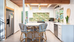 Una cocina moderna con una isla central, dos sillas de mimbre, un refrigerador y un ventanal con vistas tropicales.