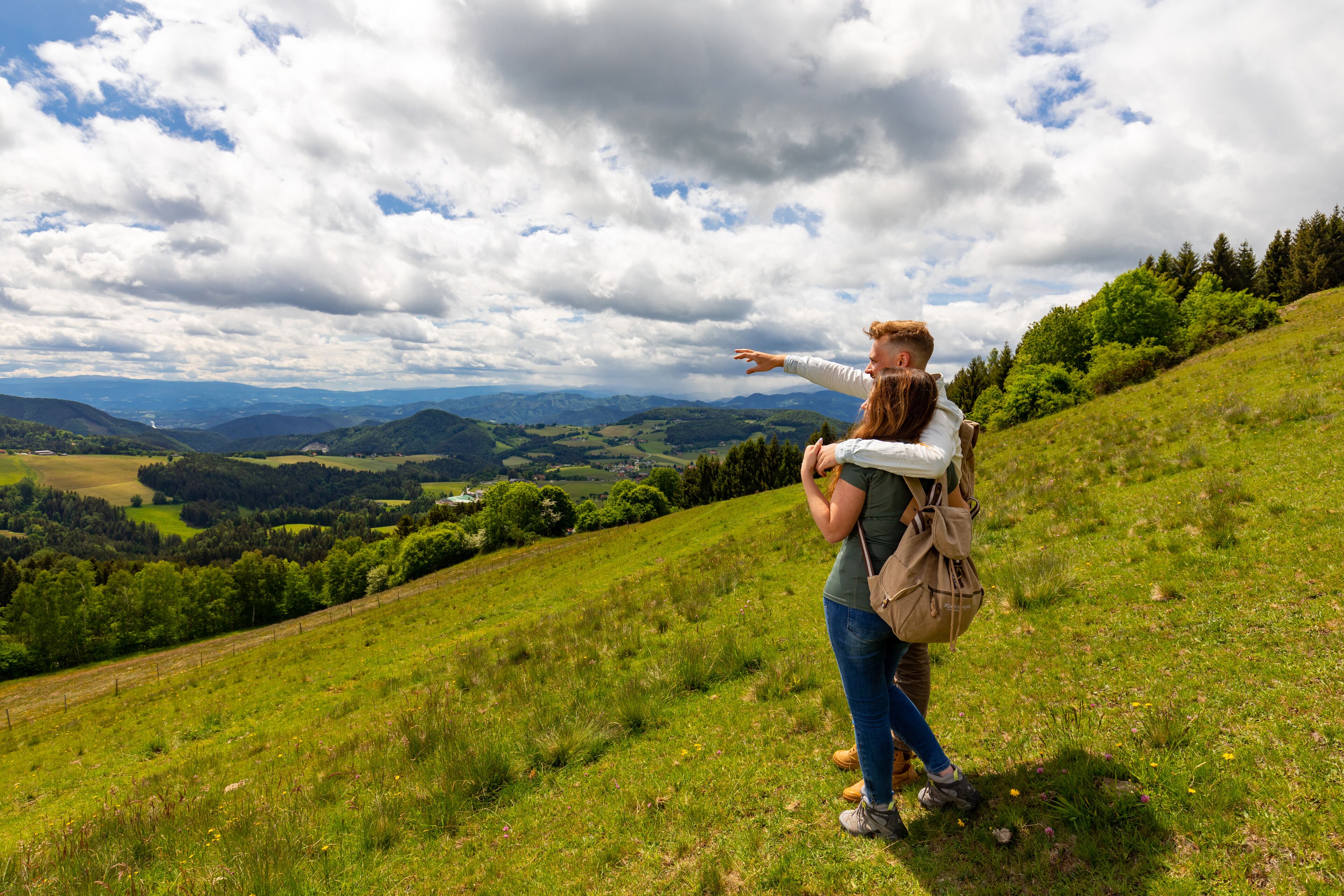 Una pareja parada en una colina cubierta de hierba con una vista panorámica de colinas ondulantes y un cielo parcialmente nublado.