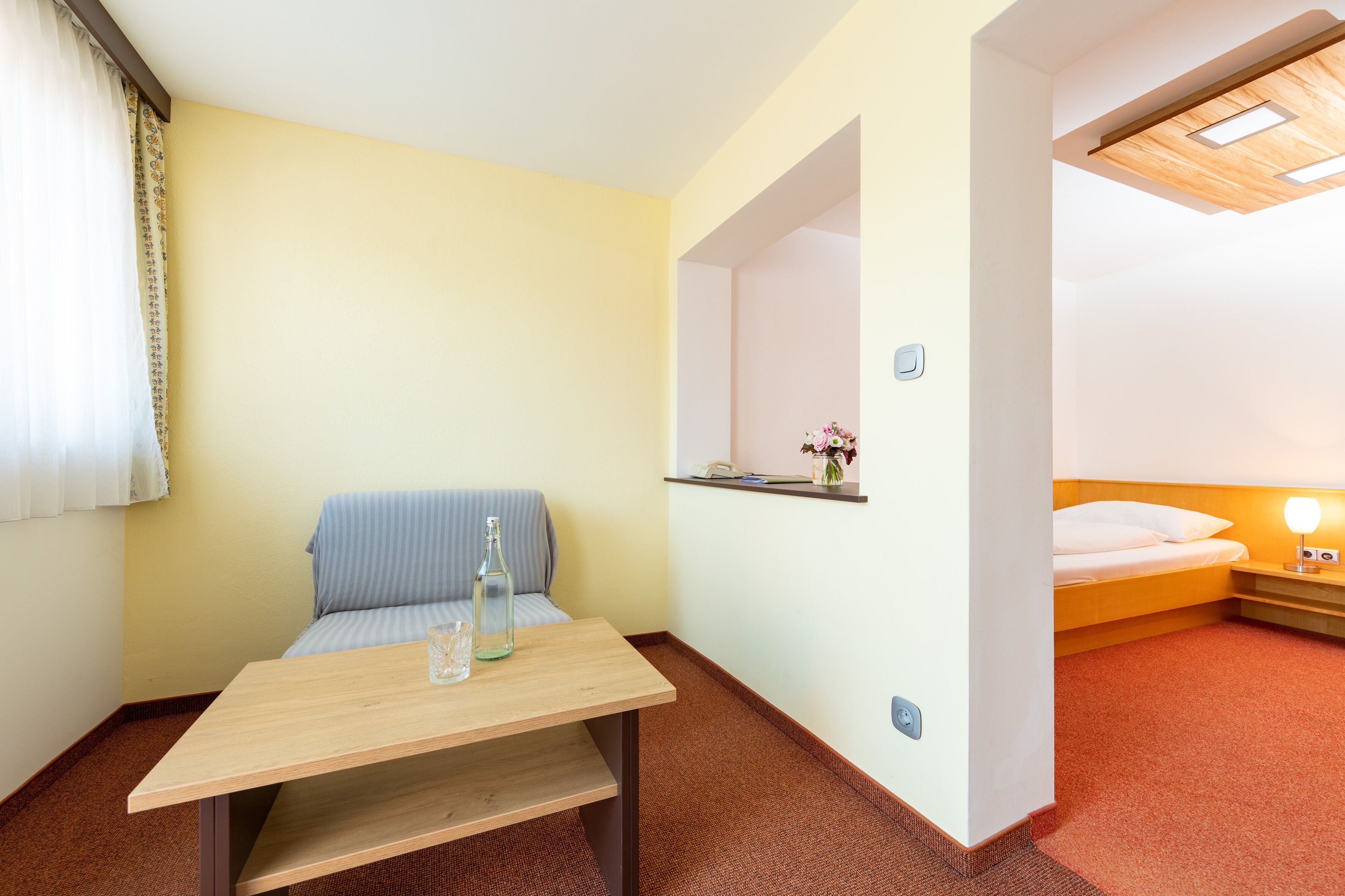Hotelkamer met een bed, nachtkastje met fles en glas, spiegel en raam met gordijnen.