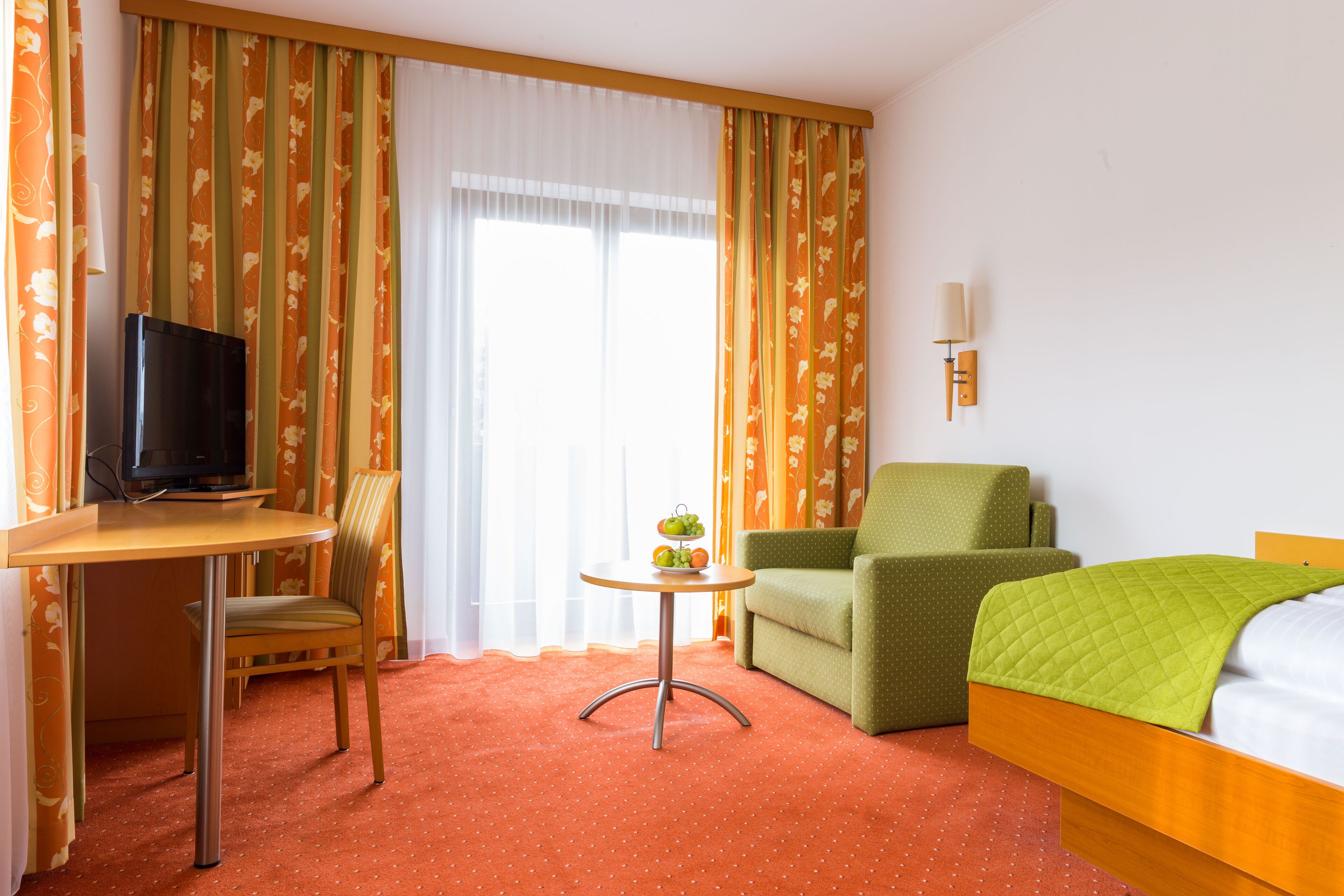 A hotel room with a bed, a desk with a television, a chair, a small table with fruit, and curtains.