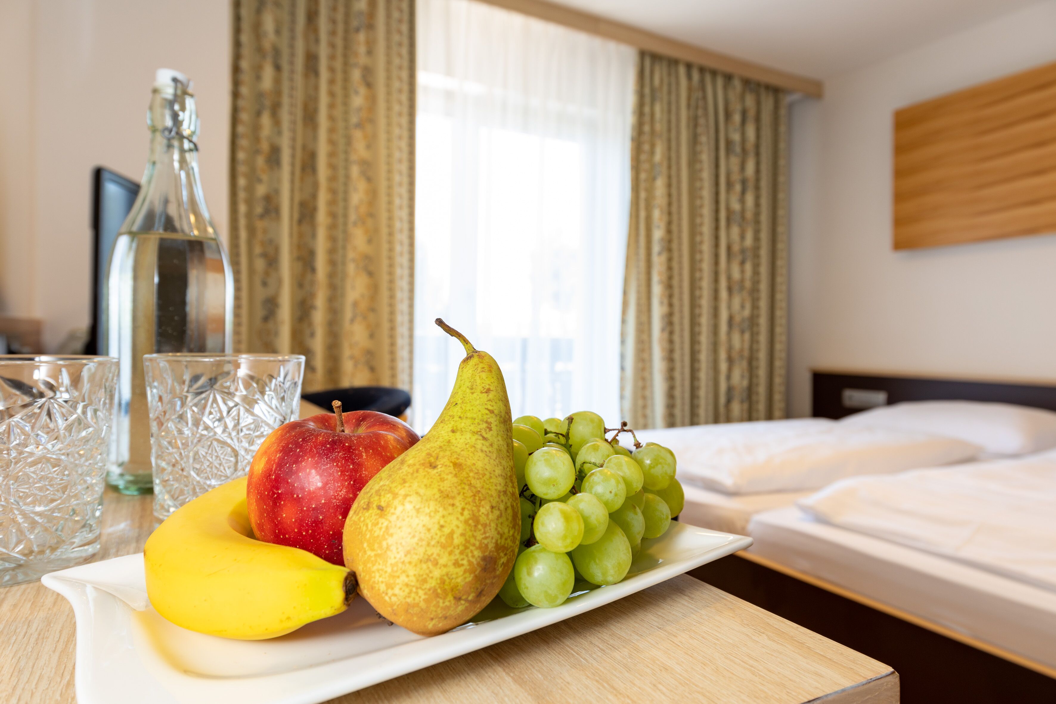 A fruit platter with an apple, pear, grapes, and banana on a white plate in a hotel room with a bed and curtains in the background.