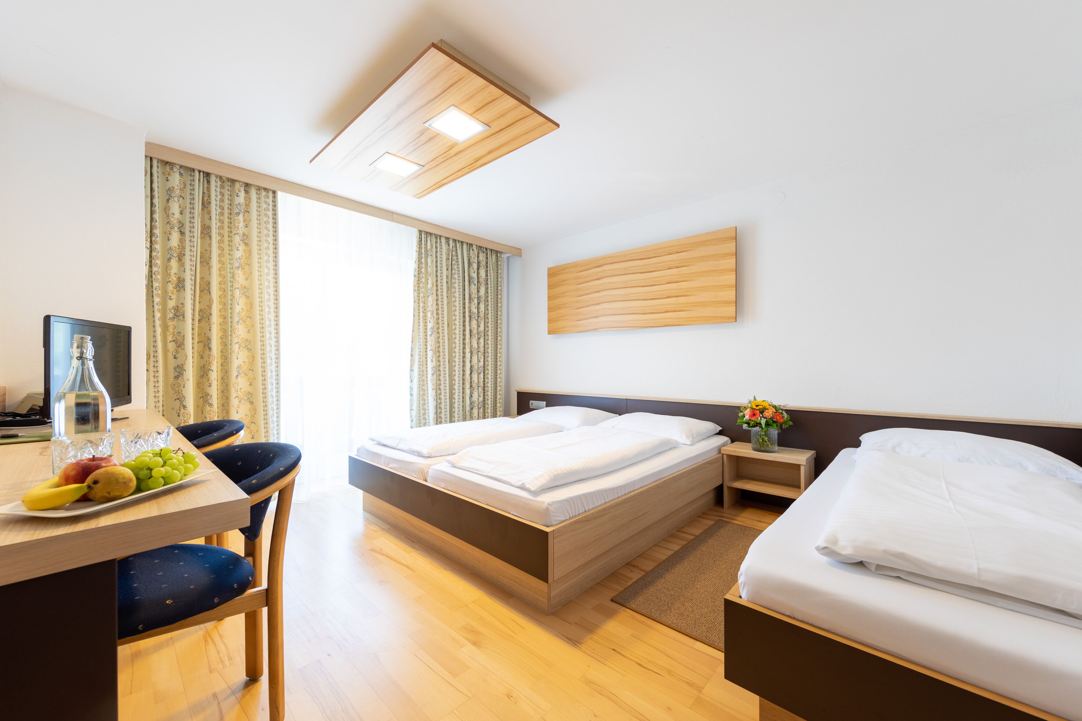 A hotel room with a wooden bed, a desk with a laptop, and a dining table with fruits.