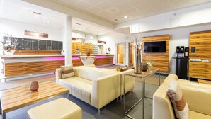 A modern hotel lobby with a reception desk, leather seating, a coffee machine, and a TV mounted on the wall.