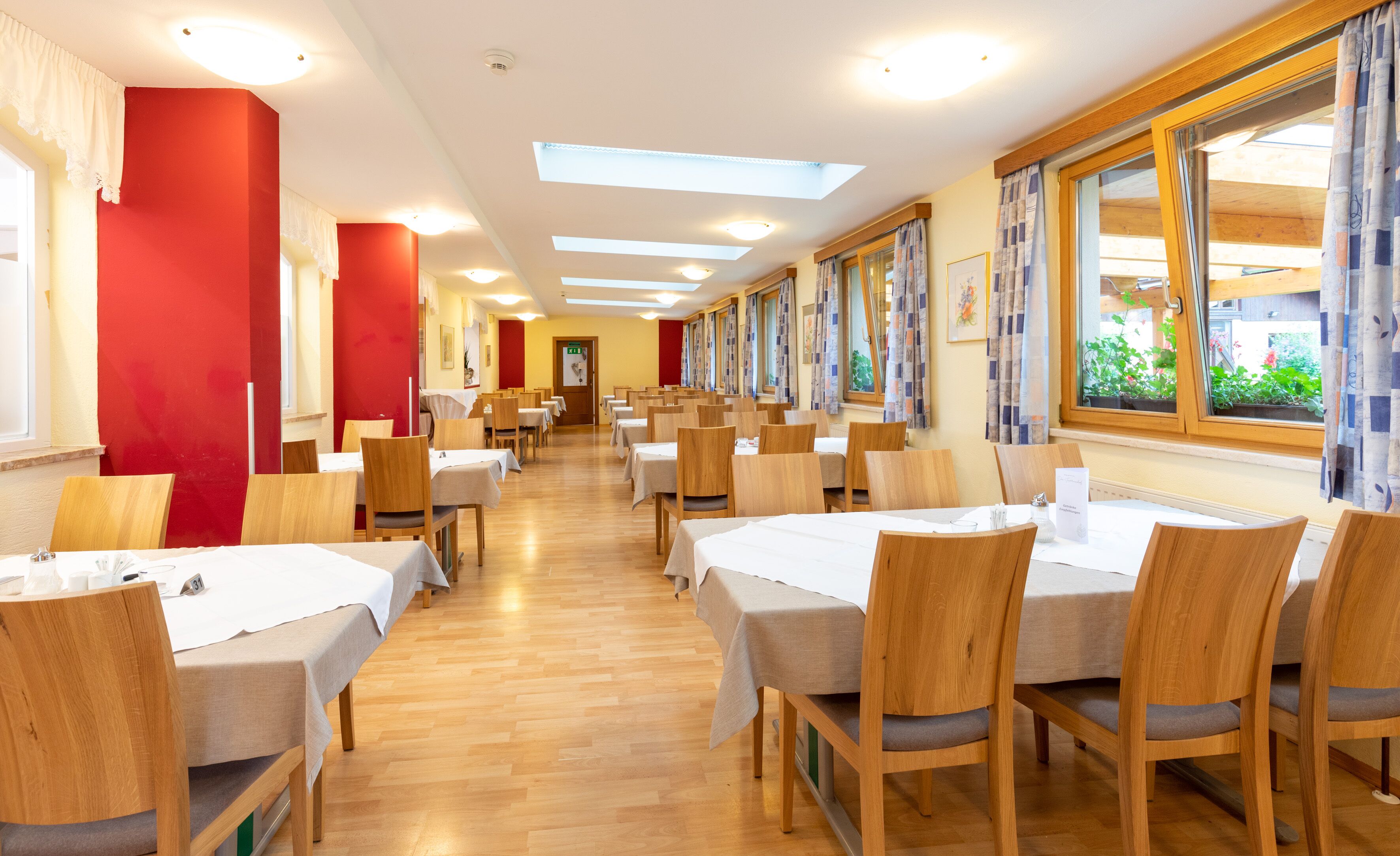 A hotel dining area with multiple tables and chairs, large windows with curtains, and a wooden floor.