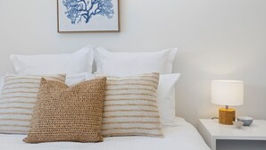 Un lit bien fait, avec des oreillers blancs et un plaid marron texturé, une œuvre d’art représentant un arbre, encadrée, accrochée au mur, et une table de chevet avec une lampe et un bol.