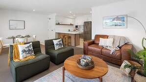 Un salon moderne avec un canapé en cuir marron, deux fauteuils verts et une table basse en bois. On aperçoit une cuisine à l’arrière-plan, avec des meubles blancs et des appareils électroménagers en acier inoxydable.
