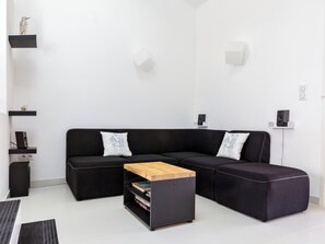 A modern living room with a black sectional sofa, a wooden coffee table, and a black ottoman. There are two white cushions on the sofa, and a small shelf with a cat figurine.