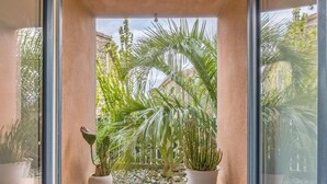 A window with two potted plants, a stone wall, and a view of a garden with palm trees and a building in the background. - Opuntia Location en Méditerranée (Escales)