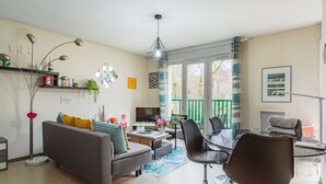 Un salon moderne avec un canapé gris, des chaises noires, une table basse en verre et un téléviseur. Il y a un balcon avec une rambarde verte et une vue sur les arbres.
