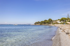 A beach with clear water, a pebble shore, and buildings in the background. - House on the beachfront with 4 bedrooms, 2 bathrooms and barbecue (Alcúdia)