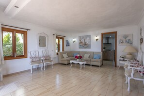A spacious living room with a beige sofa, white armchairs, and a coffee table with flowers. There is a large mirror, artwork on the walls, and a view of the kitchen through an open door. - House on the beachfront with 4 bedrooms, 2 bathrooms and barbecue (Alcúdia)