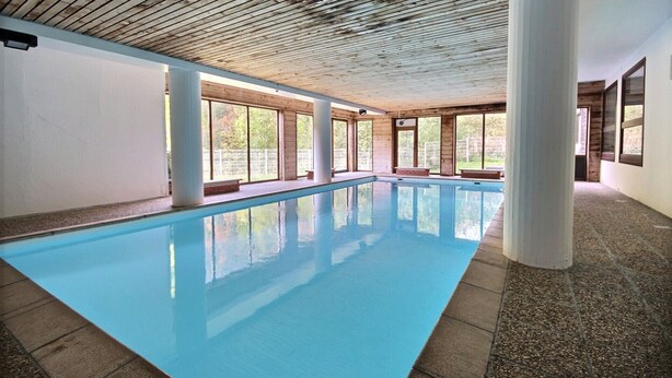 Una piscina interior con agua azul cristalina, vigas de madera en el techo, ventanas amplias y suelo de baldosas.