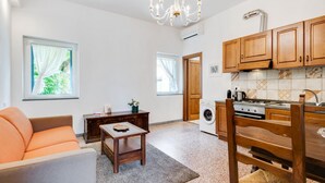 A living room with a sofa, a table, a chandelier, and a kitchen with wooden cabinets and a washing machine.