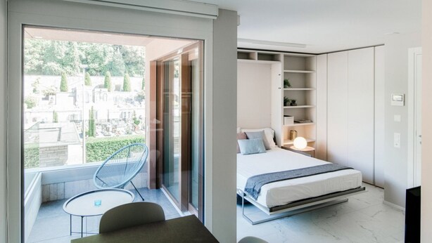 A modern bedroom with a bed, bedside table, and a view through a glass door to an outdoor area with a chair and table. - Calm Studio near the center (WHITE STUDIO 8) (Locarno)