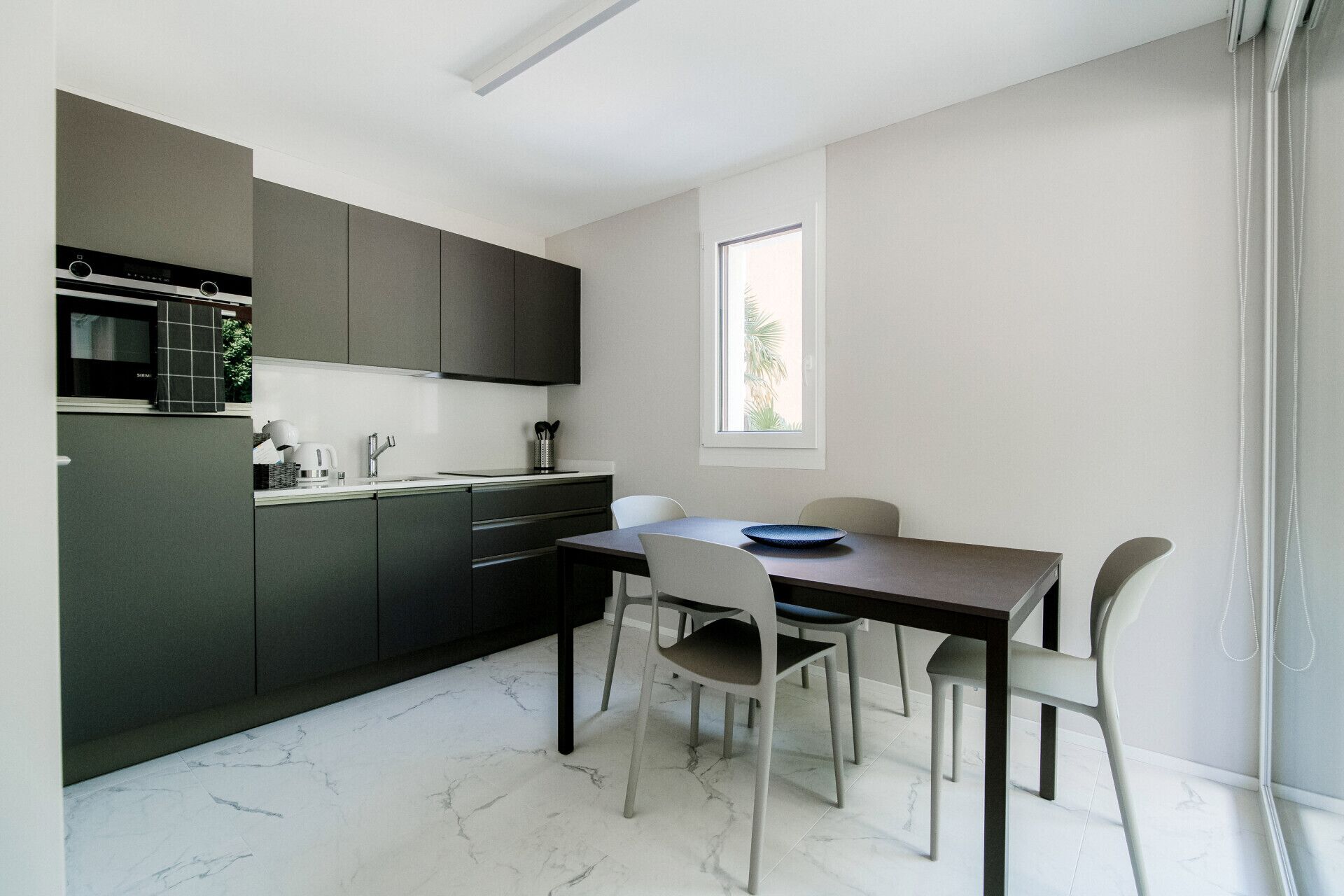 Une cuisine moderne avec des meubles de couleur sombre, un four encastré et une fenêtre qui laisse entrer la lumière naturelle.