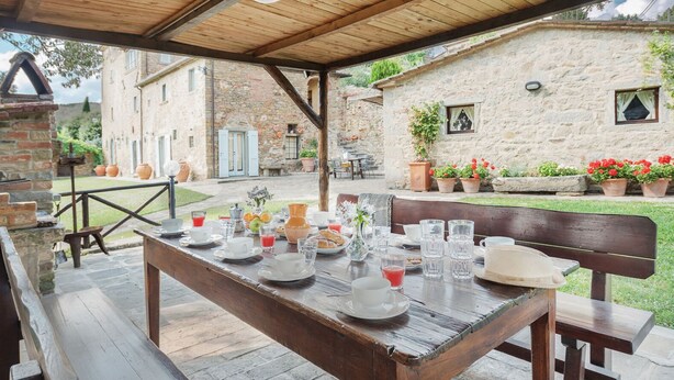 An outdoor dining area with a rustic wooden table set for breakfast, surrounded by stone buildings and potted plants. - Elks Tower (Toscana)