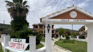 La entrada de Villa Corfiota, con un cartel, un camino y vistas de la villa y la vegetación circundante.