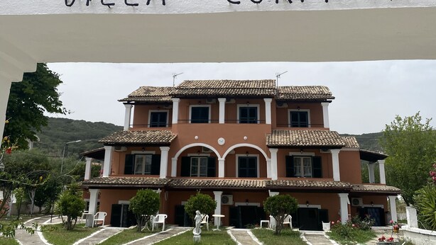 Villa Corfiota con un llamativo cartel de entrada, un edificio de varios pisos con tejado de tejas y un jardín ajardinado con senderos.