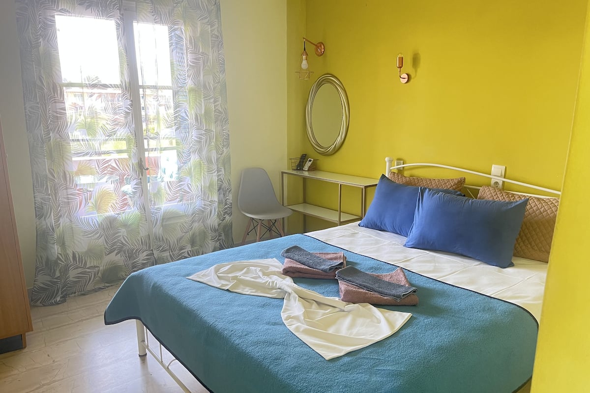 Een net opgemaakt bed met blauw-witte beddengoed, een stoel, een nachtkastje en een spiegel aan een gele muur.