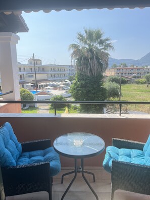 A balcony with two wicker chairs and a glass table, overlooking a pool and buildings.