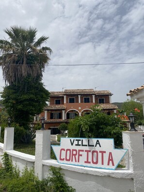 Une villa nommée « VILLA CORFIOTA », dotée d’un toit traditionnel en tuiles, entourée d’une végétation luxuriante et d’un palmier.