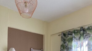 Una habitación con pared beige, un pequeño escritorio blanco con cajones, un espejo, una lámpara de pared y una ventana con cortina de hojas tropicales.