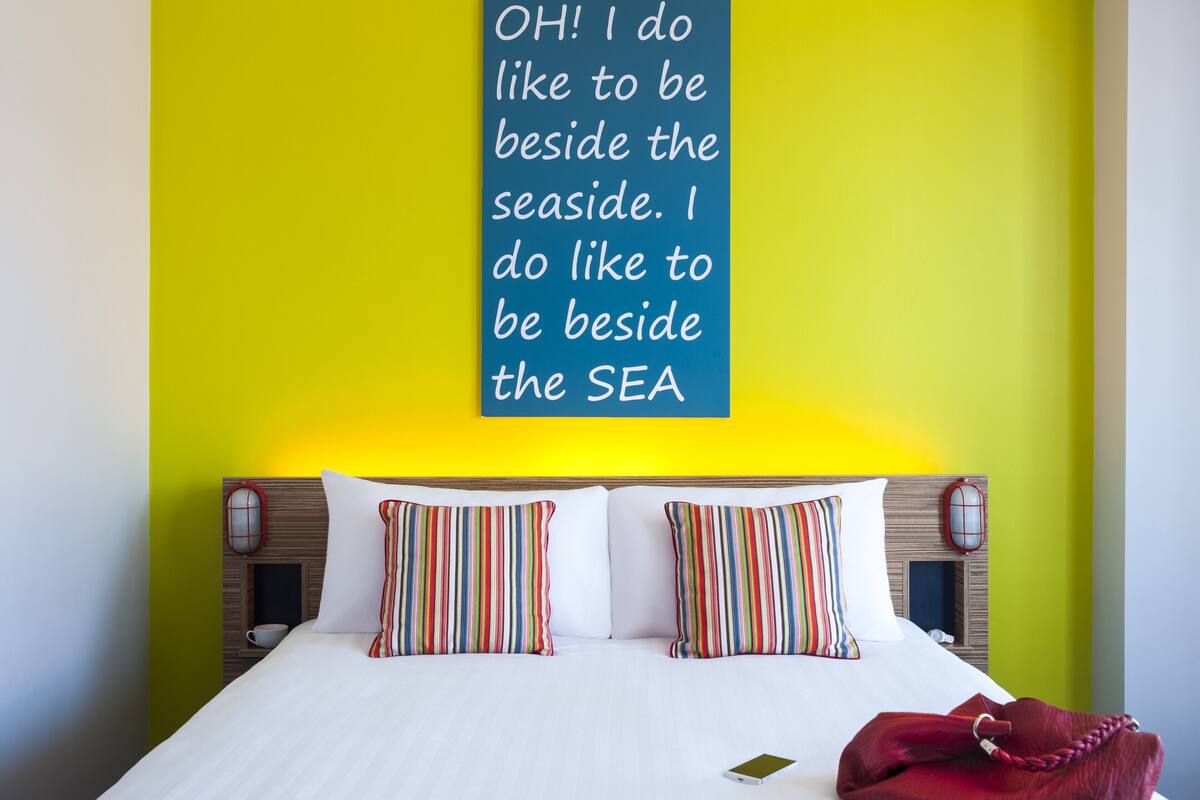 A hotel room with a bed, two striped pillows, a red throw blanket, and a wall with a motivational quote.