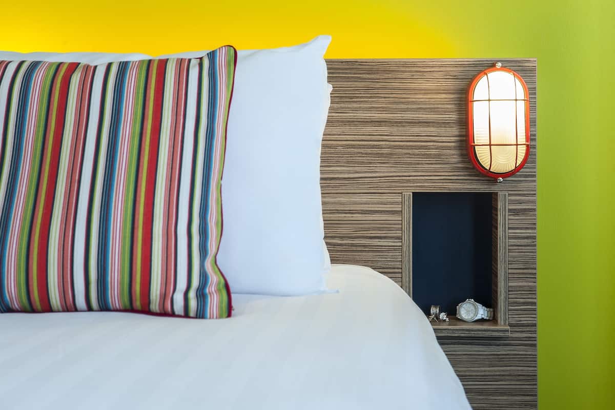 A bed with striped and white pillows, a wooden headboard, a red-framed mirror, and a watch on the mirror.