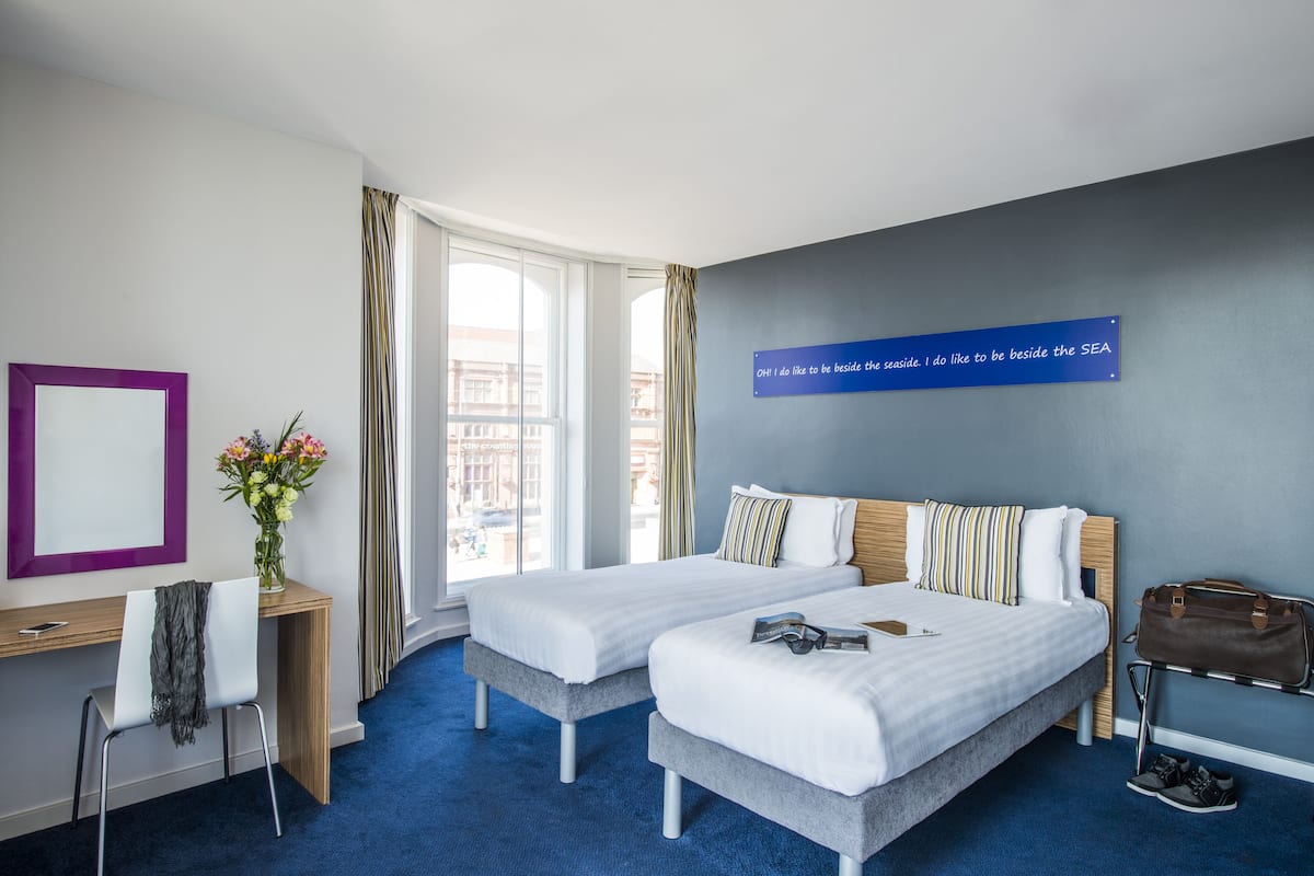 A hotel room with two beds, a desk, a chair, a vase with flowers, a handbag, and a sign on the wall.