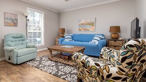 Un salon avec un canapé bleu, un fauteuil bleu clair, un fauteuil à motifs, une table basse et un ventilateur de plafond.
