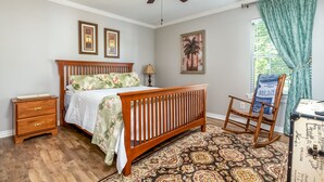 Une chambre à coucher avec un lit en bois, une table de chevet, un fauteuil à bascule, un ventilateur de plafond et une fenêtre avec des rideaux.