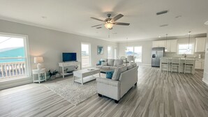Ein geräumiges Wohnzimmer mit einem großen Sofa, einem Couchtisch und einem Fernseher. Es gibt einen Balkon mit Blick, eine Küchenzeile mit weißen Schränken und einen Deckenventilator.