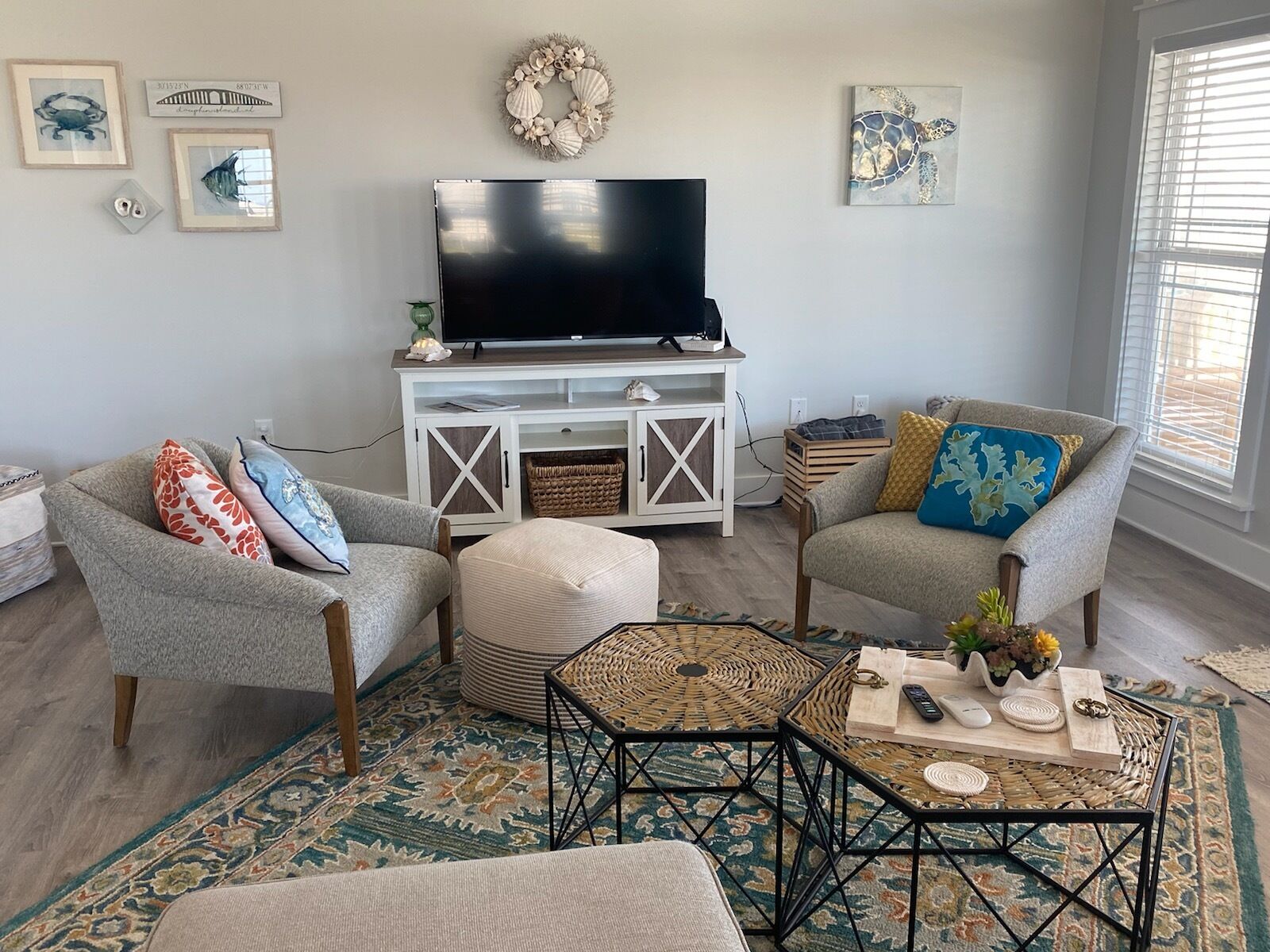 Un salon avec deux fauteuils gris, un meuble de rangement blanc intégrant un téléviseur à écran plat, une table basse en fibres tressées et un tapis à motifs.