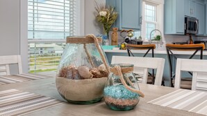 Een eettafel met twee glazen potten met decoratieve spullen, een raam met luxaflex en een keuken met blauwe kastjes en een magnetron.