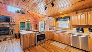 A wooden cabin-style kitchen with a stone fireplace, wooden cabinets, a stainless steel dishwasher, and a large window. - A Backdrop Dream - 3BR/3BA Log Home, Hot Tub, Firepit (Maggie Valley)