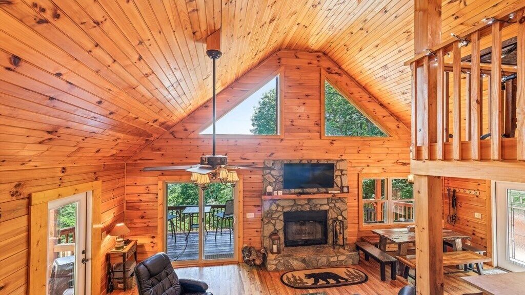 A log cabin interior with a stone fireplace, wooden ceiling, and a dining area.