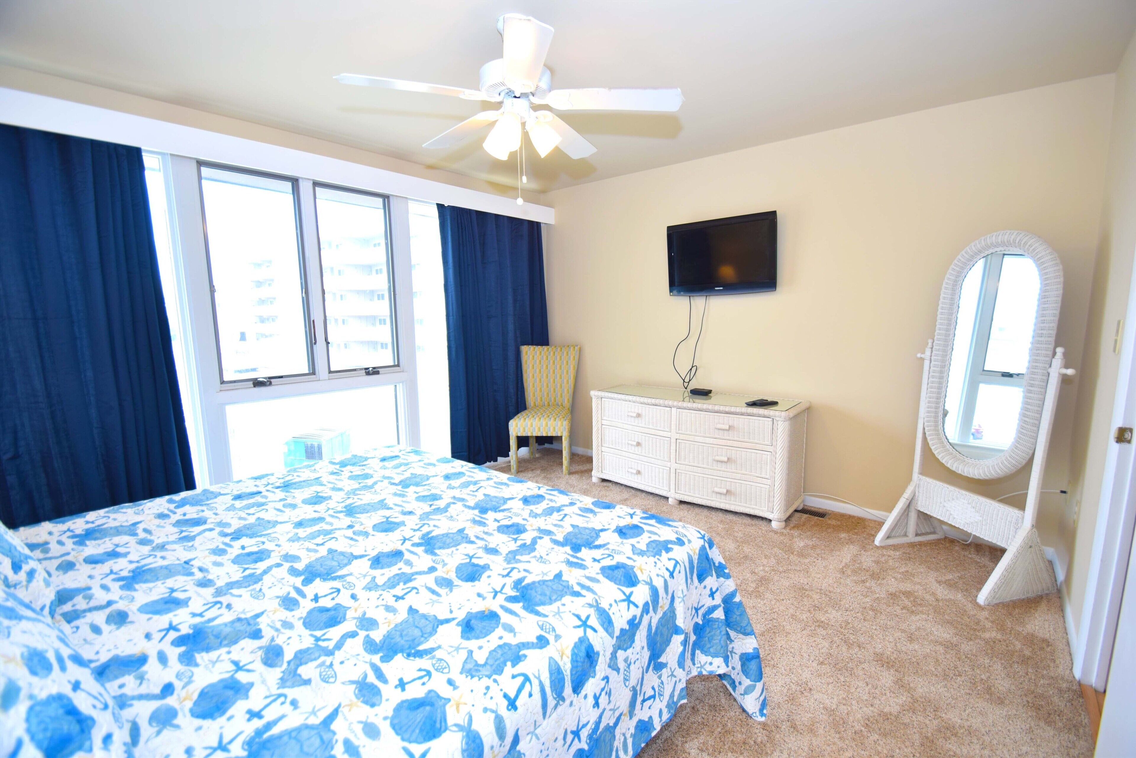 A bedroom with a bed, a dresser, a television, a ceiling fan, and a window with curtains.