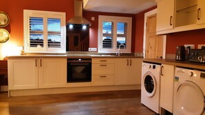 A compact kitchen with white cabinets, a built-in oven, a washing machine, and a window with blinds. - Seaside Cottage in Renvyle (Connemara)