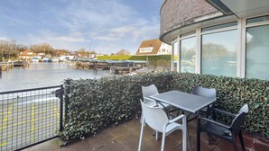 Une terrasse avec une table et des chaises donnant sur un canal, avec une haie et un bâtiment aux grandes fenêtres en arrière-plan.