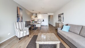 Una sala de estar moderna con un sofá, un sillón y una mesa de centro. Se ve una cocina al fondo.