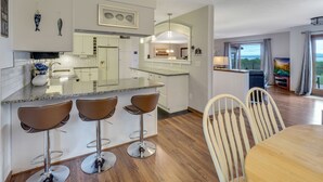 A modern kitchen with a central island, bar stools, and a dining area with a wooden table and chairs. - Beautiful views of Little Traverse Bay and a spacious wraparound deck (Petoskey)