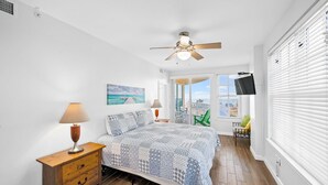 Une chambre à coucher comprenant un lit, une table de chevet, un ventilateur de plafond, une fenêtre avec des stores et une vue sur la ville.