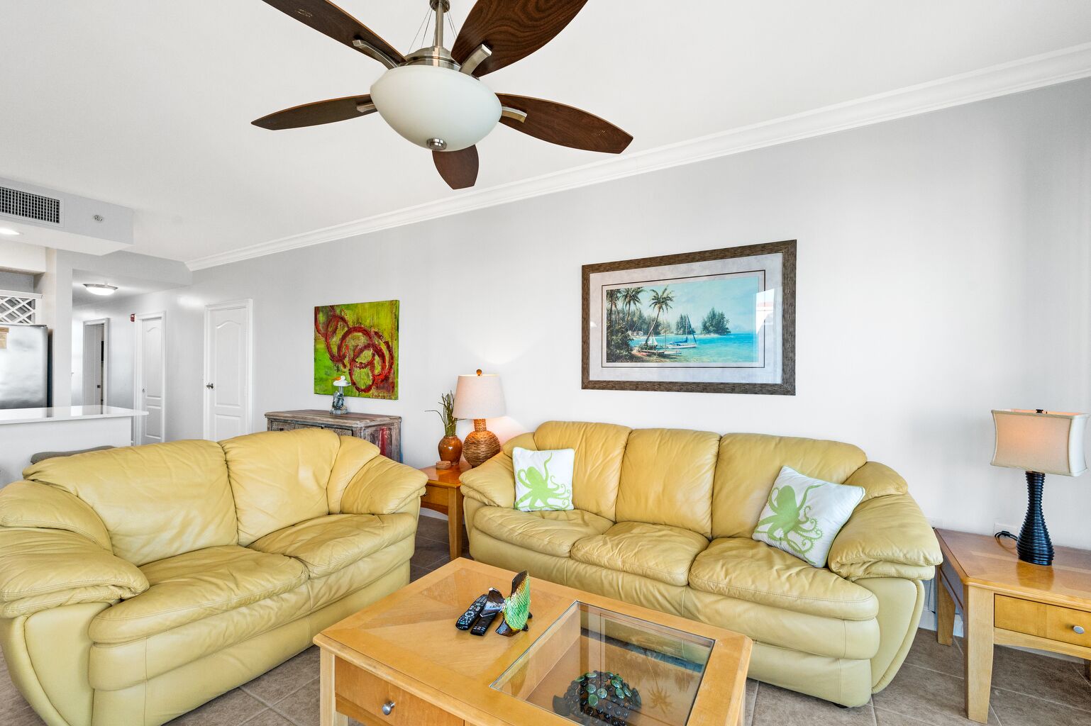 A living room with two yellow leather sofas, a glass coffee table, a ceiling fan, a framed picture, and a lamp on a wooden side table.