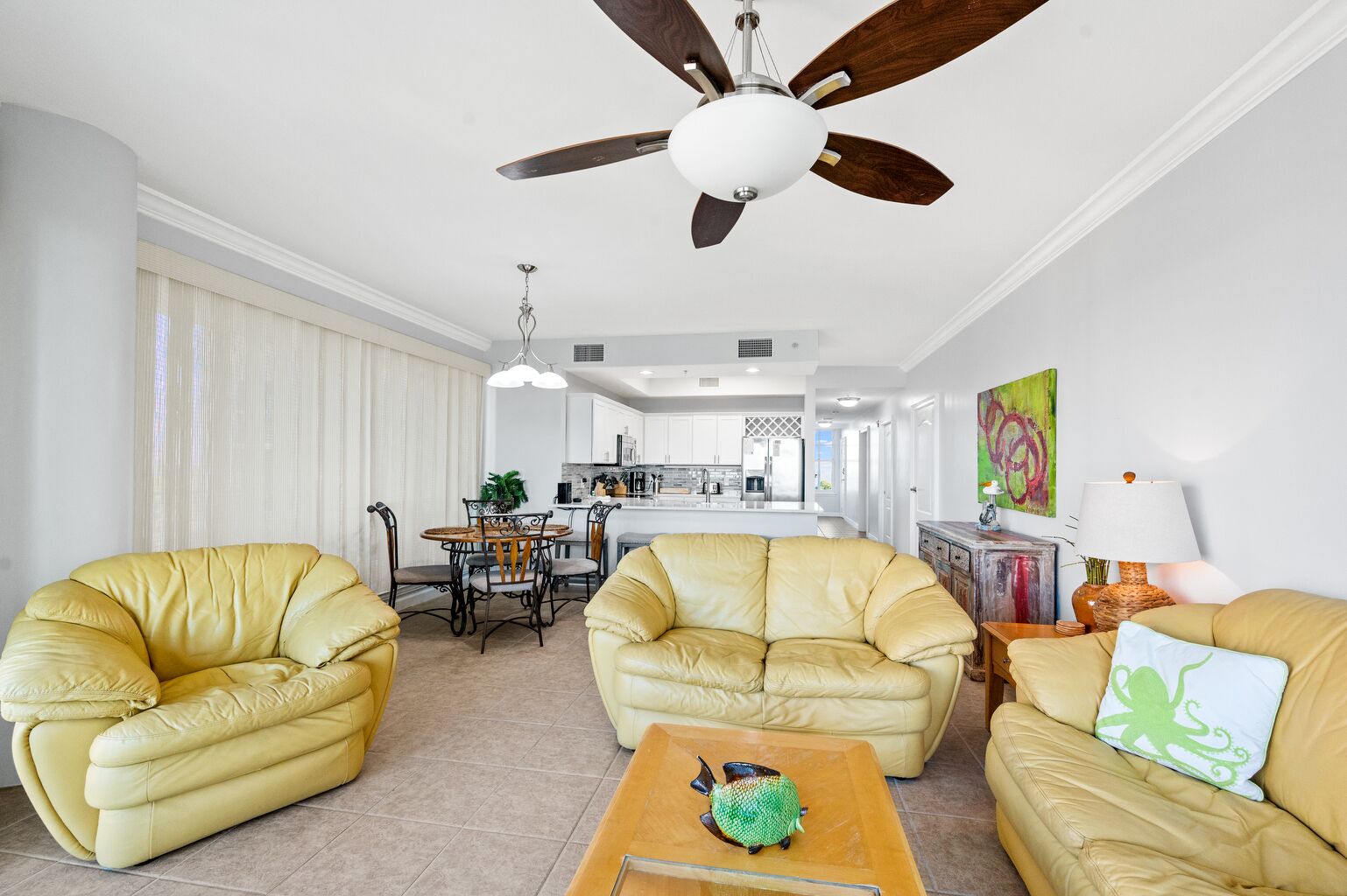 A living room with a ceiling fan, a dining area with a table and chairs, and a kitchen in the background.