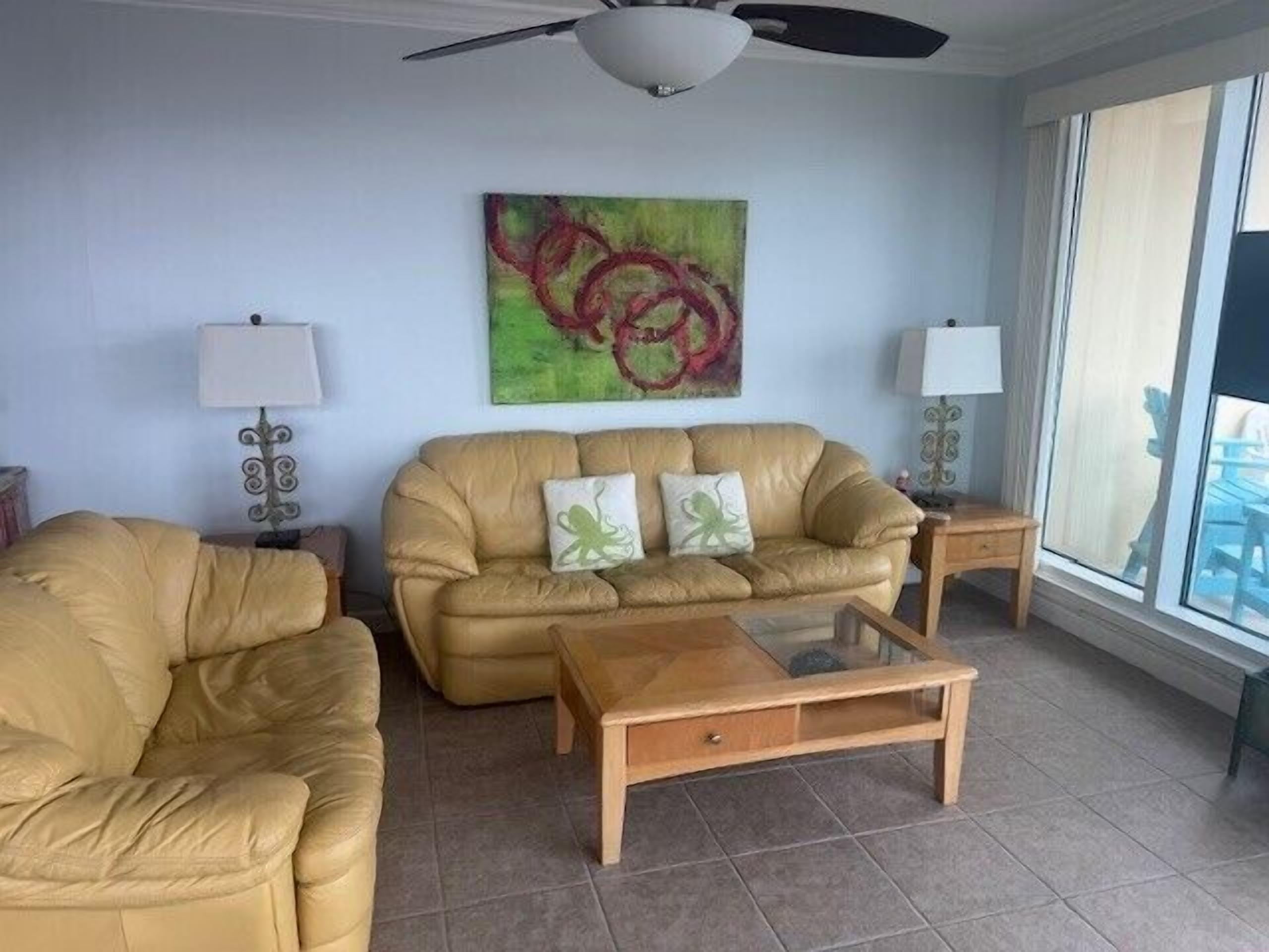 A living room with a beige sofa, a glass coffee table, and two side tables with lamps. There is a large abstract painting on the wall and a window with blinds.