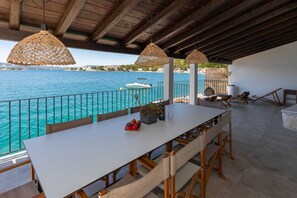 Un espacio para comer al aire libre con vista al mar, una mesa blanca y lámparas colgantes de mimbre.