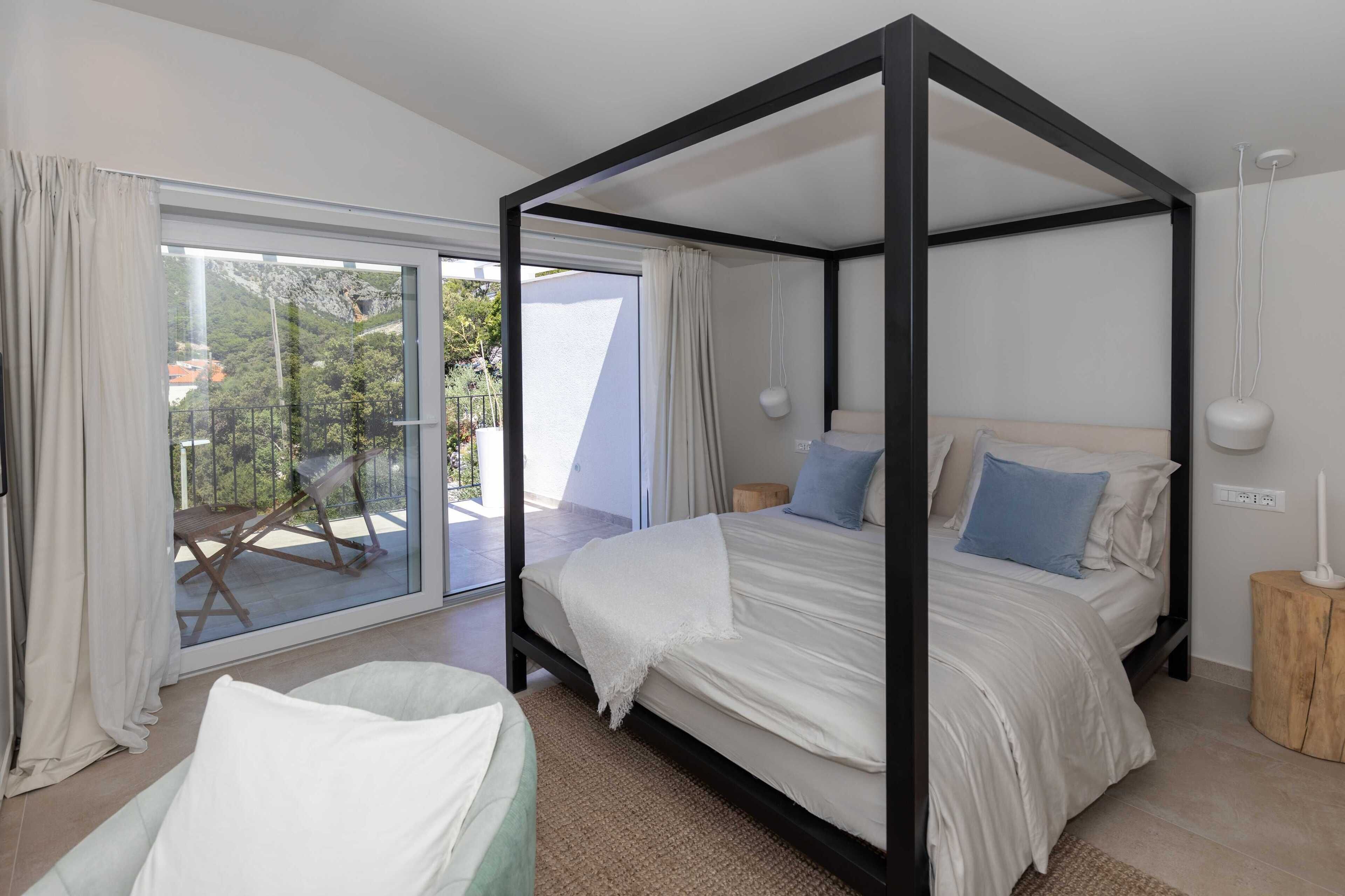 A four-poster bed with white bedding, a wooden nightstand, and a view of a balcony with outdoor furniture.