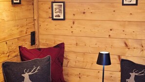 Ein gemütliches Zimmer mit Holzwänden, einem weichen roten Kissen, zwei Kissen mit Reh-Motiv, einer schwarzen Lampe und einem dekorativen Nest mit Kerzen.