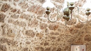 Une pièce avec un mur en pierre, un lit recouvert de draps blancs, une table ornée de bougies et des chaises recouvertes d’un tissu léger et transparent.